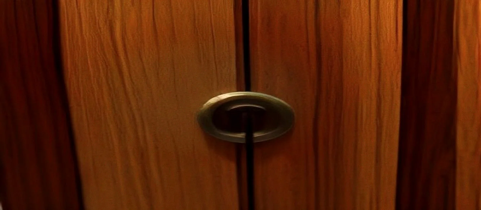 A brass keyhole escutcheon set into the edge of a dark teak wardrobe door, the metal dulled and scratched.