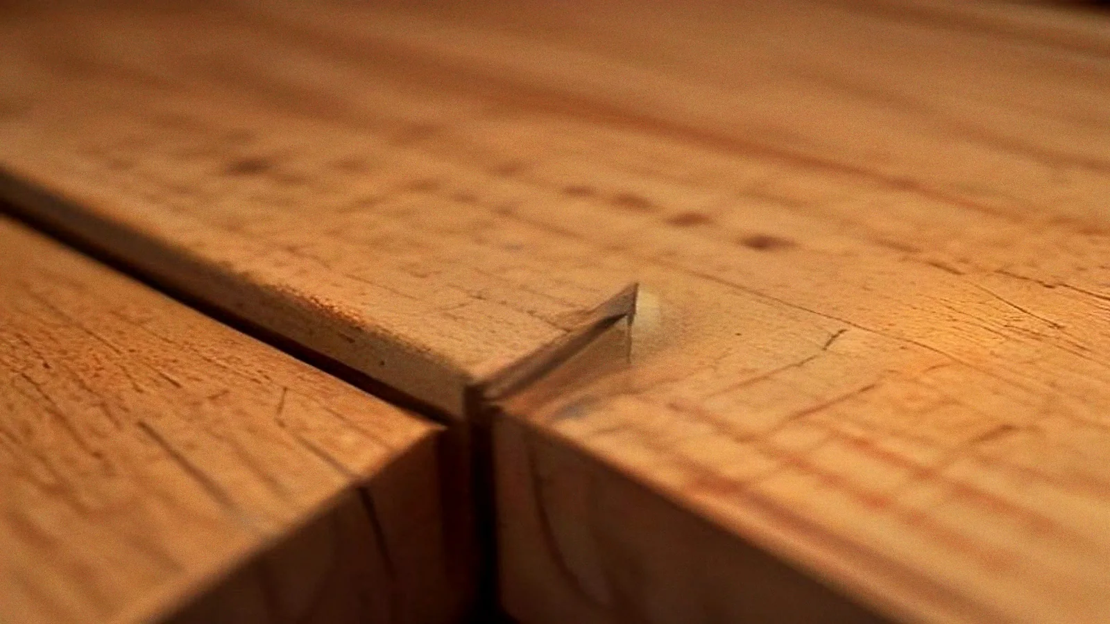 An extreme close-up of a mitred corner on a teak table, the grain running in two directions either side of a hairline gap along the joint.