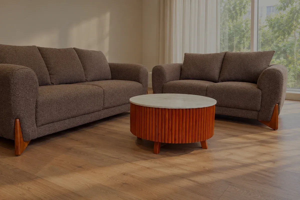 A brown textured-weave sofa and loveseat with curved arms on a pale oak floor, a linen curtain half drawn across the window behind.