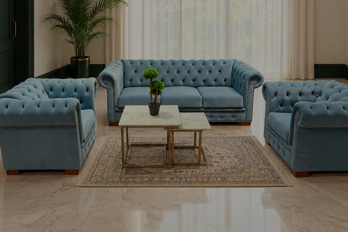 A powder-blue deep-buttoned sofa and two armchairs in a tall room, sheer curtains across a full-height window and a pale marble floor.