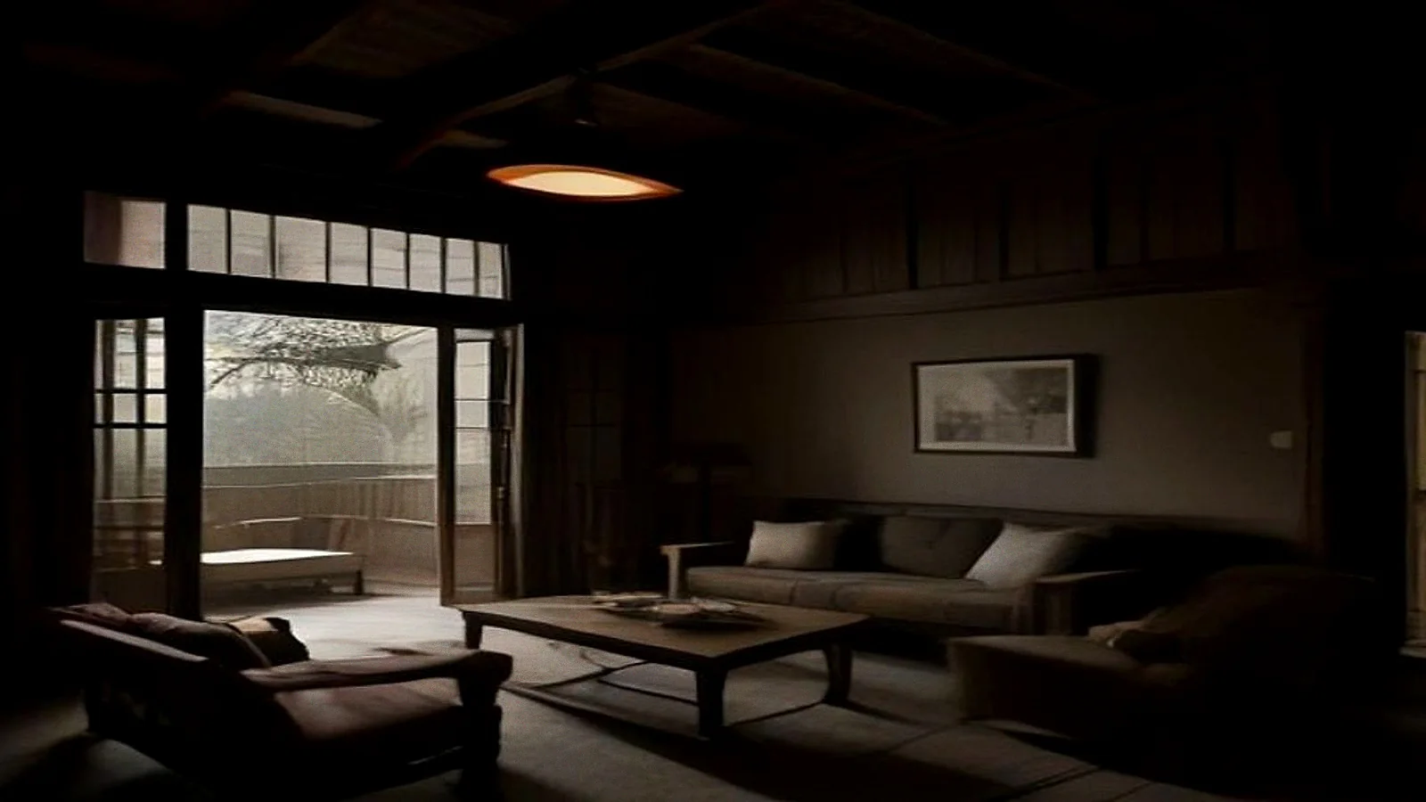 A living room at night with one lamp lit over a low teak sofa, the far end of the room in darkness and the doors open onto unlit greenery.