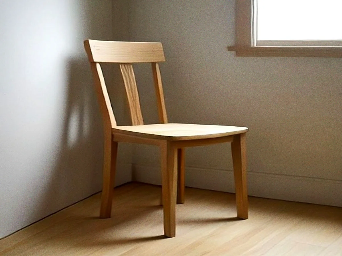 An unfinished chair in bare pale timber standing against a plain wall, no seat and no polish, the joints visible.